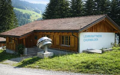 Ein Holzgebaeude mit Beschriftung steht in alpiner Landschaft bei Churwalden (7075, Kanton Graubuenden) und zeigt ein Lehrkraftwerk.
