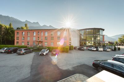 Vor den Bergen steht in Maienfeld (7304, Kanton Graubuenden) ein Hotelgebäude mit Glasfassade und Parkplätzen, während die Sonne über den Gipfeln aufgeht.