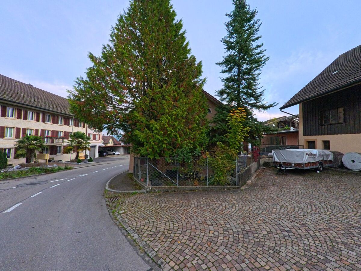 In Tägerig (5522, Kanton Aargau) verläuft eine schmale Strasse zwischen Wohnhäusern, während rechts ein gepflasterter Hof mit einem abgedeckten Anhänger und hohen Bäumen liegt.