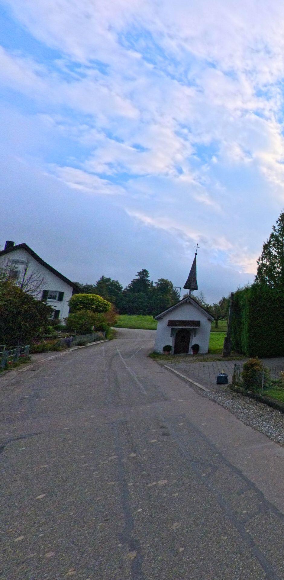 In Tägerig (5522, Kanton Aargau) führt eine ruhige Strasse an einer kleinen Kapelle vorbei, hinter der sich eine Wiese und ein bewaldeter Hang ausbreiten.
