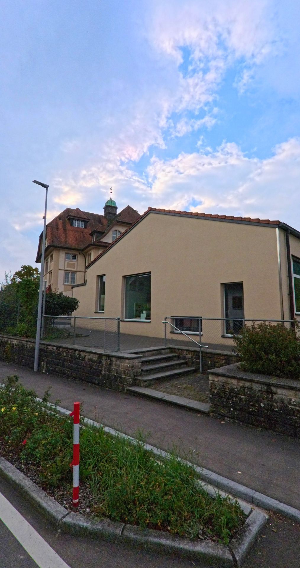 In Tägerig (5522, Kanton Aargau) steht ein modernes, helles Gebäude vor einem älteren Haus mit markantem Turm, während ein Gehweg und eine kleine bepflanzte Verkehrsinsel im Vordergrund liegen.