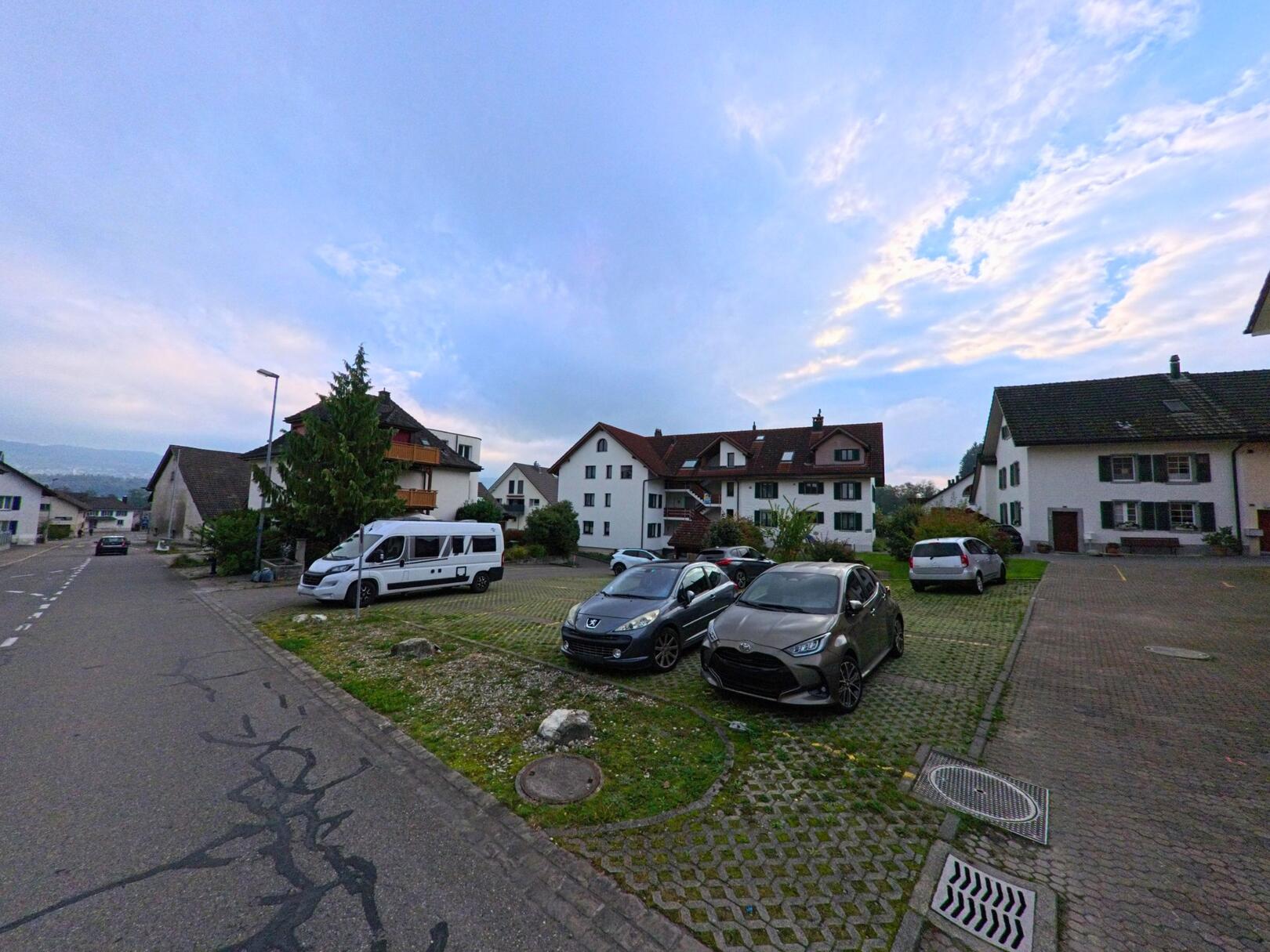 In einem Wohnquartier von Tägerig (5522, Kanton Aargau) stehen mehrere Fahrzeuge auf einem kleinen Parkplatz zwischen gepflegten Häusern unter einem leicht bewölkten Abendhimmel.