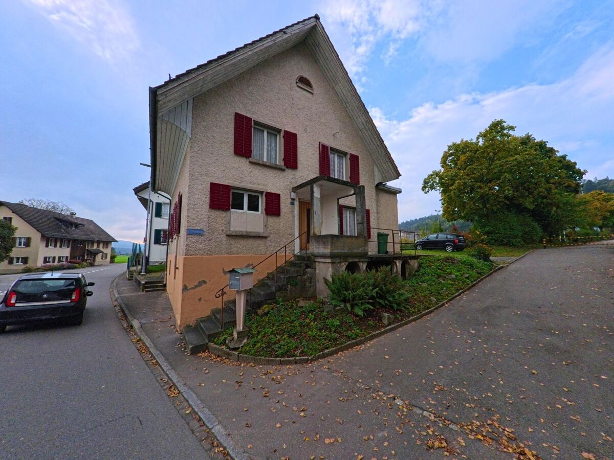In Tägerig (5522, Kanton Aargau) steht ein älteres Wohnhaus mit roten Fensterläden an einer Strassenkreuzung, umgeben von Bäumen und weiteren Gebäuden.