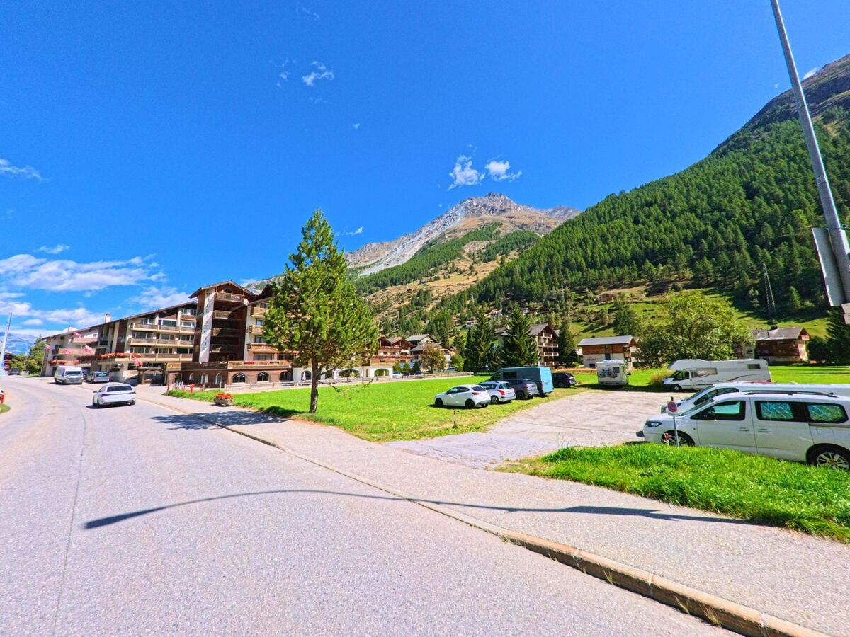 Am Ortsrand von Täsch (3929, Kanton Wallis) verlaufen Strasse und Parkflächen vor Hotels und Wohnhäusern, dahinter erheben sich bewaldete Berghänge.