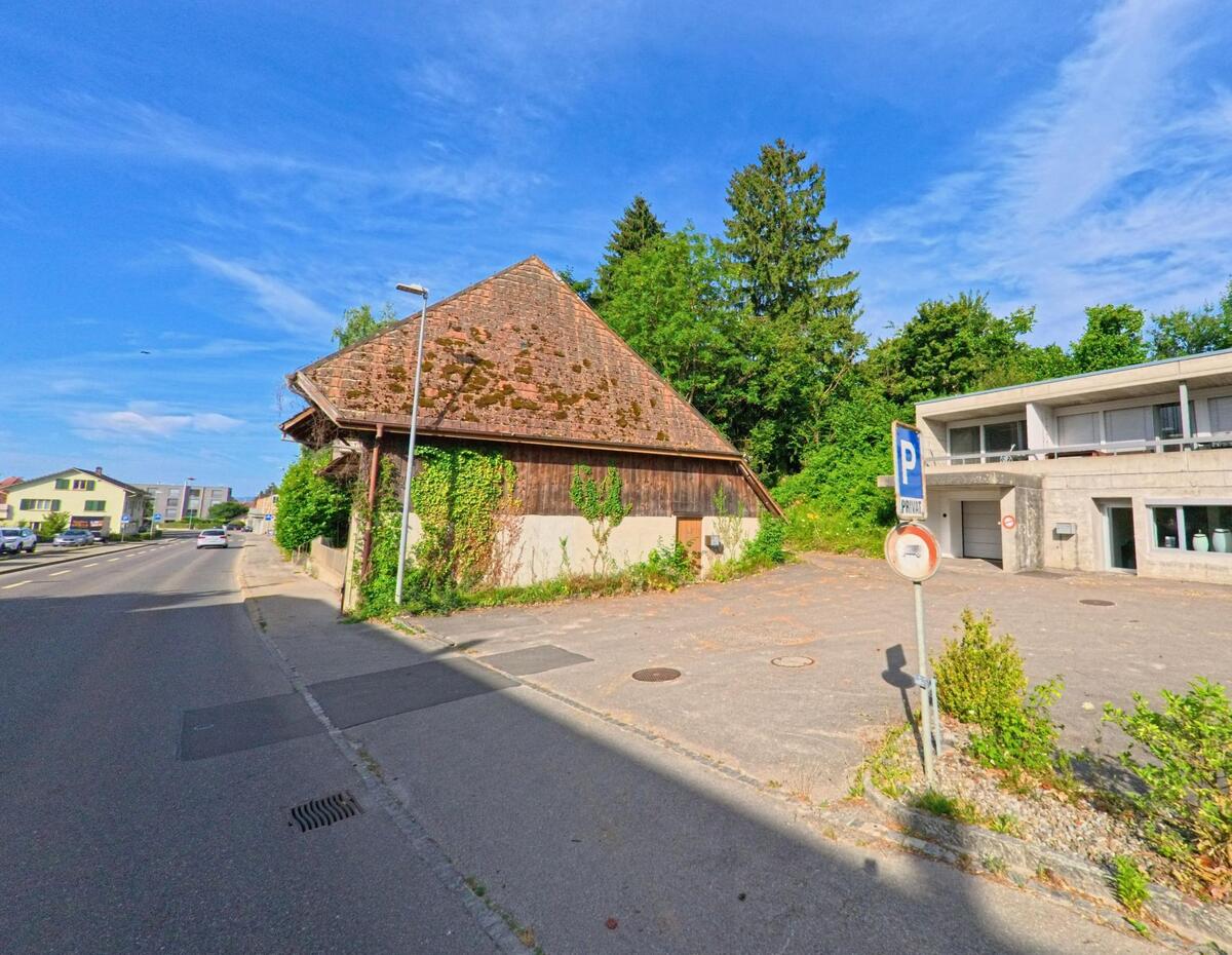 In Täuffelen (2575, Kanton Bern) steht ein altes, moosbewachsenes Holzgebäude neben einem modernen Flachbau mit Parkplätzen an einer Hauptstrasse.
