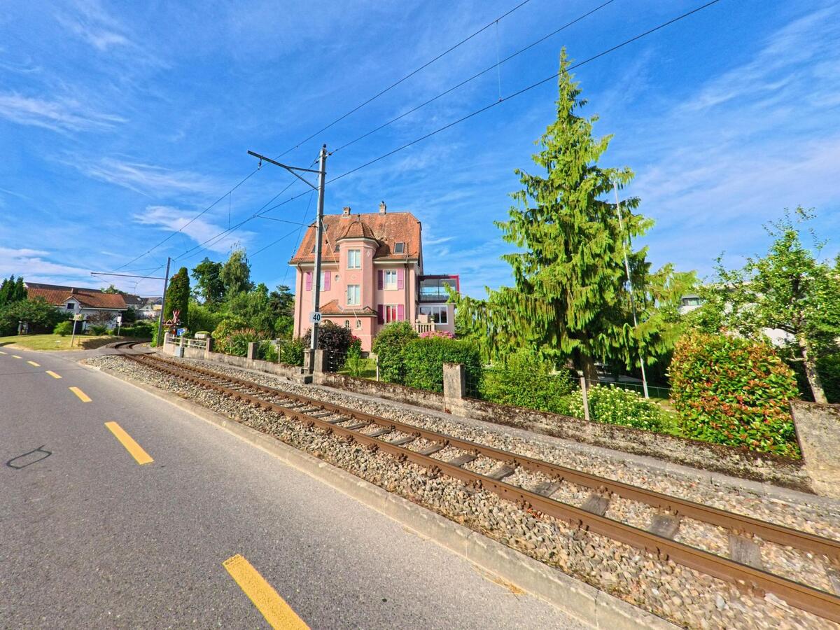 In Täuffelen (2575, Kanton Bern) verläuft eine Bahnlinie direkt an einem auffällig rosafarbenen Haus mit gepflegtem Garten und Nadelbäumen vorbei.