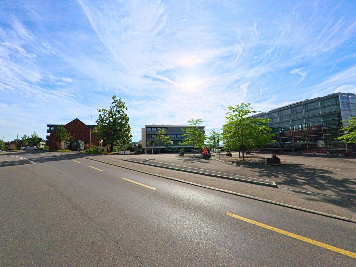 In Täuffelen (2575, Kanton Bern) öffnet sich ein grosser asphaltierter Platz mit jungen Bäumen und modernen Gebäuden aus Glas und Beton im Hintergrund.