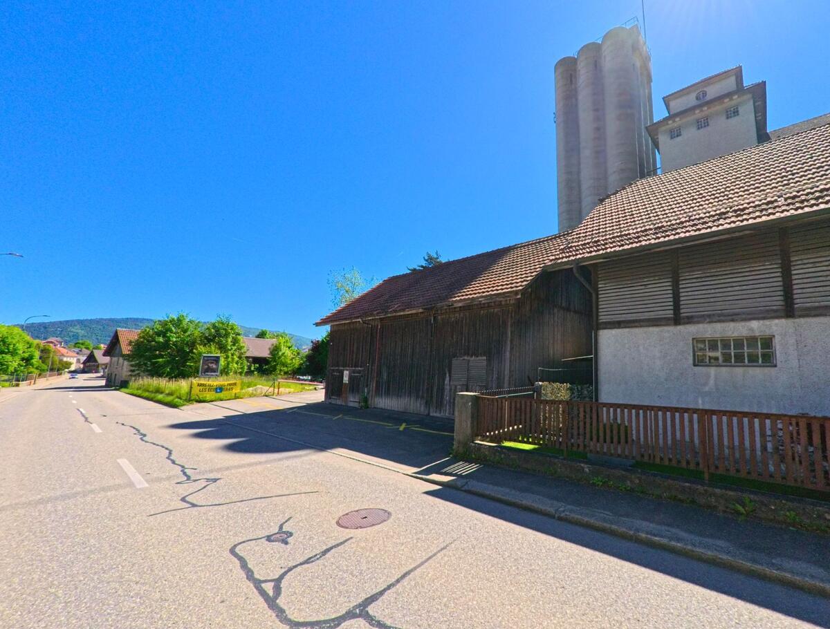 In Tavannes (2710, Kanton Bern) führt eine Strasse entlang eines alten Industriegebäudes mit Silos, das von grünen Bäumen umgeben ist.