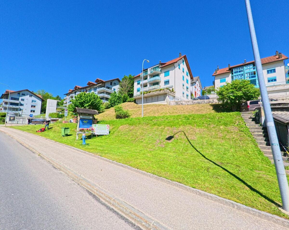 In Tavannes (2710, Kanton Bern) führt eine Strasse an modernen Wohngebäuden entlang, wobei ein „Bienvenue“-Schild und eine gepflegte Grünfläche den Eingang zur Ortschaft markieren.