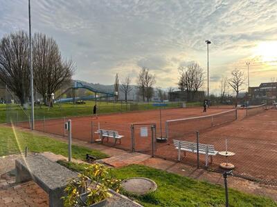 Auf der Tennisanlage in Schmerikon (8716, Kanton St. Gallen) spielen zwei Personen auf gepflegten Sandplaetzen, waehrend die Abendsonne hinter Baumen und Rutschenlandschaft sanft ueber dem Gelände steht.