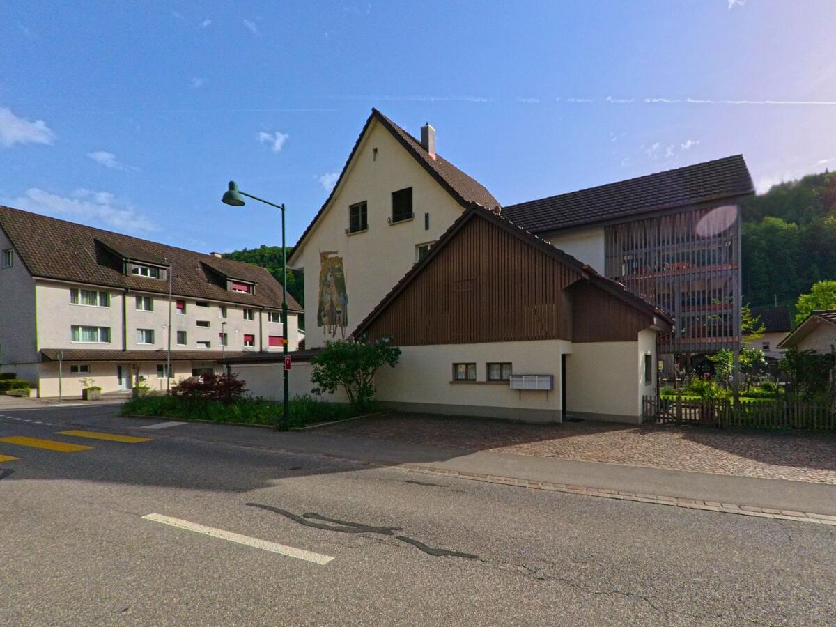 In Tecknau (4492, Kanton Basel-Landschaft) zeigt eine Fassade mit Wandmalerei ein traditionell gestaltetes Gebäude gegenüber modernen Mehrfamilienhäusern.