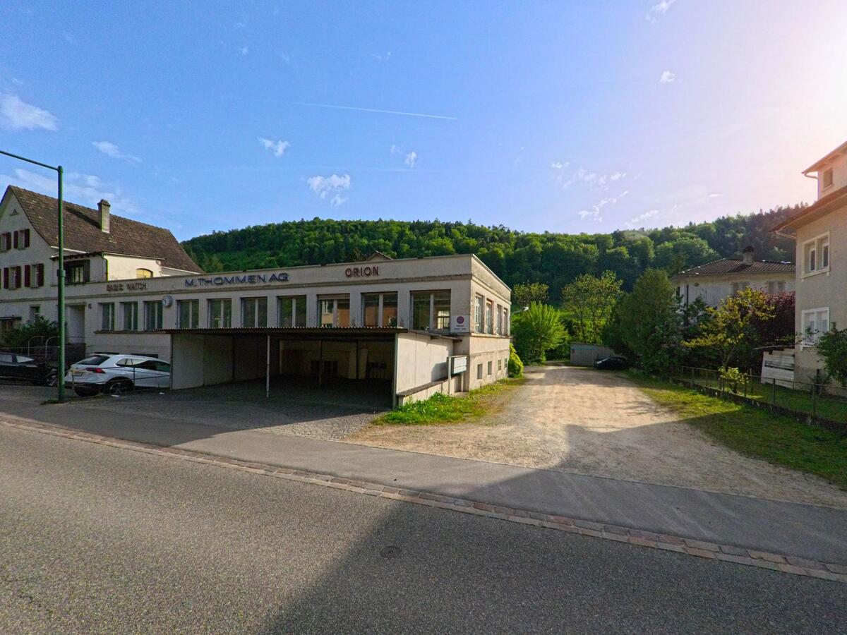 In Tecknau (4492, Kanton Basel-Landschaft) befindet sich ein flaches Gewerbegebäude mit der Aufschrift „M. Thommen AG“ und angrenzendem Kiesweg zwischen Wohnhäusern.