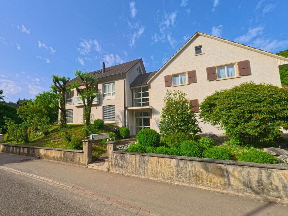 In Tecknau (4492, Kanton Basel-Landschaft) stehen moderne Mehrfamilienhäuser mit gepflegten Gärten und geschnittenen Bäumen an einer ruhigen Quartierstrasse.