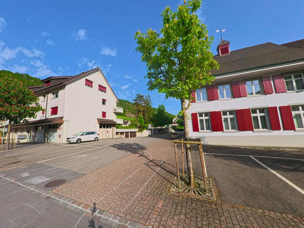 Der Platz vor dem Schulgebäude in Tecknau (4492, Kanton Basel-Landschaft) ist von gepflasterten Wegen, Parkplätzen und Bäumen umgeben.