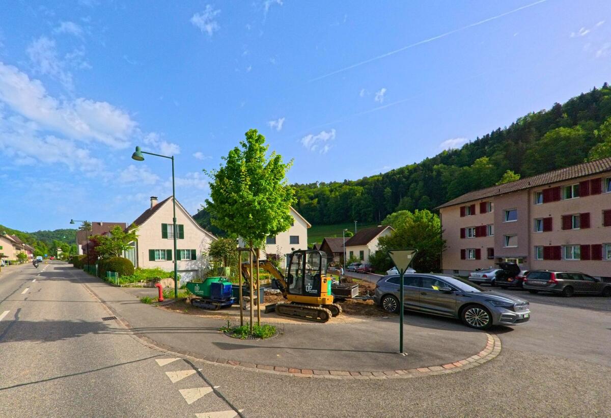 In Tecknau (4492, Kanton Basel-Landschaft) wird an einer Strassenecke zwischen Wohnhäusern eine kleine Baustelle mit Bagger betrieben.