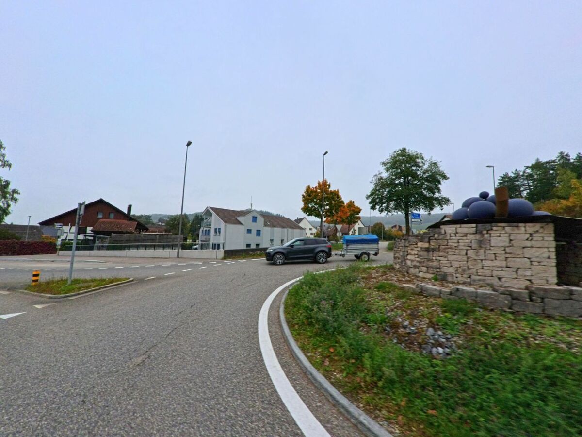 In einem Kreisverkehr von Tegerfelden (5306, Kanton Aargau) steht im Vordergrund ein Steinsockel mit runden Skulpturen, waehrend umliegende Wohnhaeuser und Baeume das Bild rahmen.