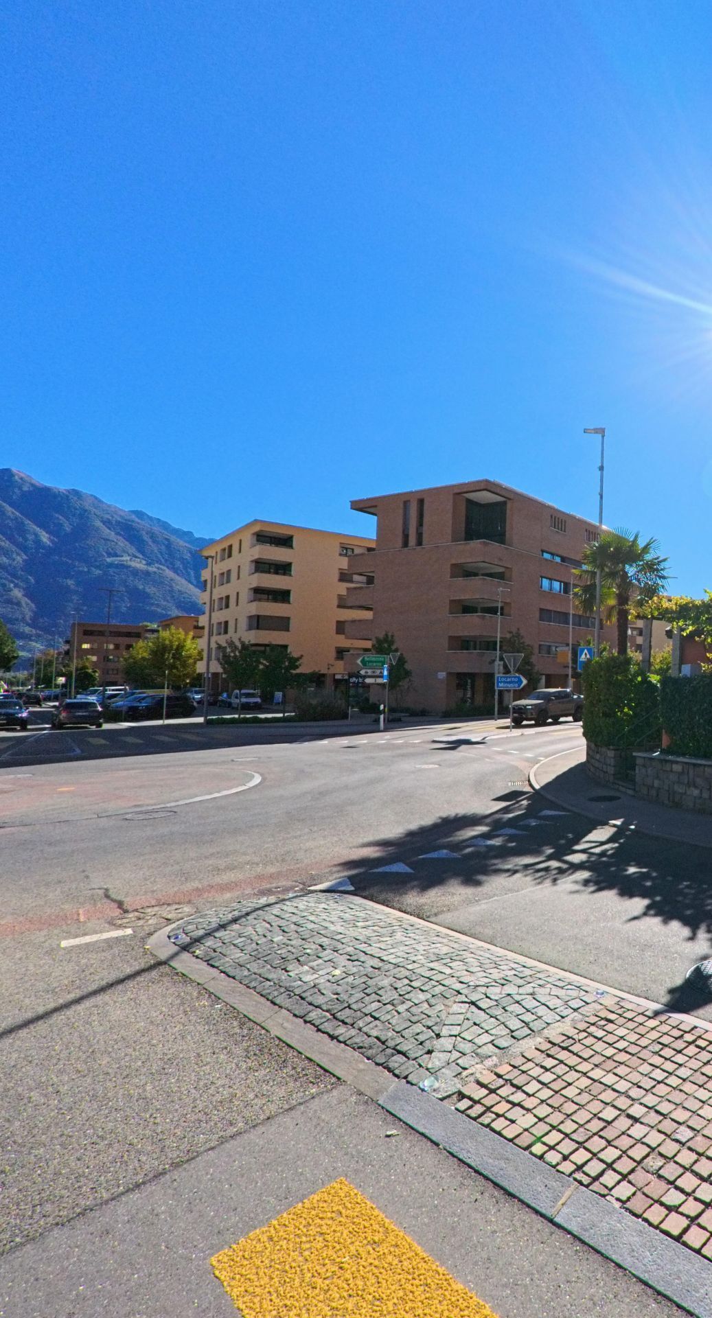 Mehrstöckige Wohnhäuser und ein Kreisverkehr prägen das Stadtbild von Tenero-Contra (6598, Kanton Tessin) mit Blick auf die umliegenden Berge.