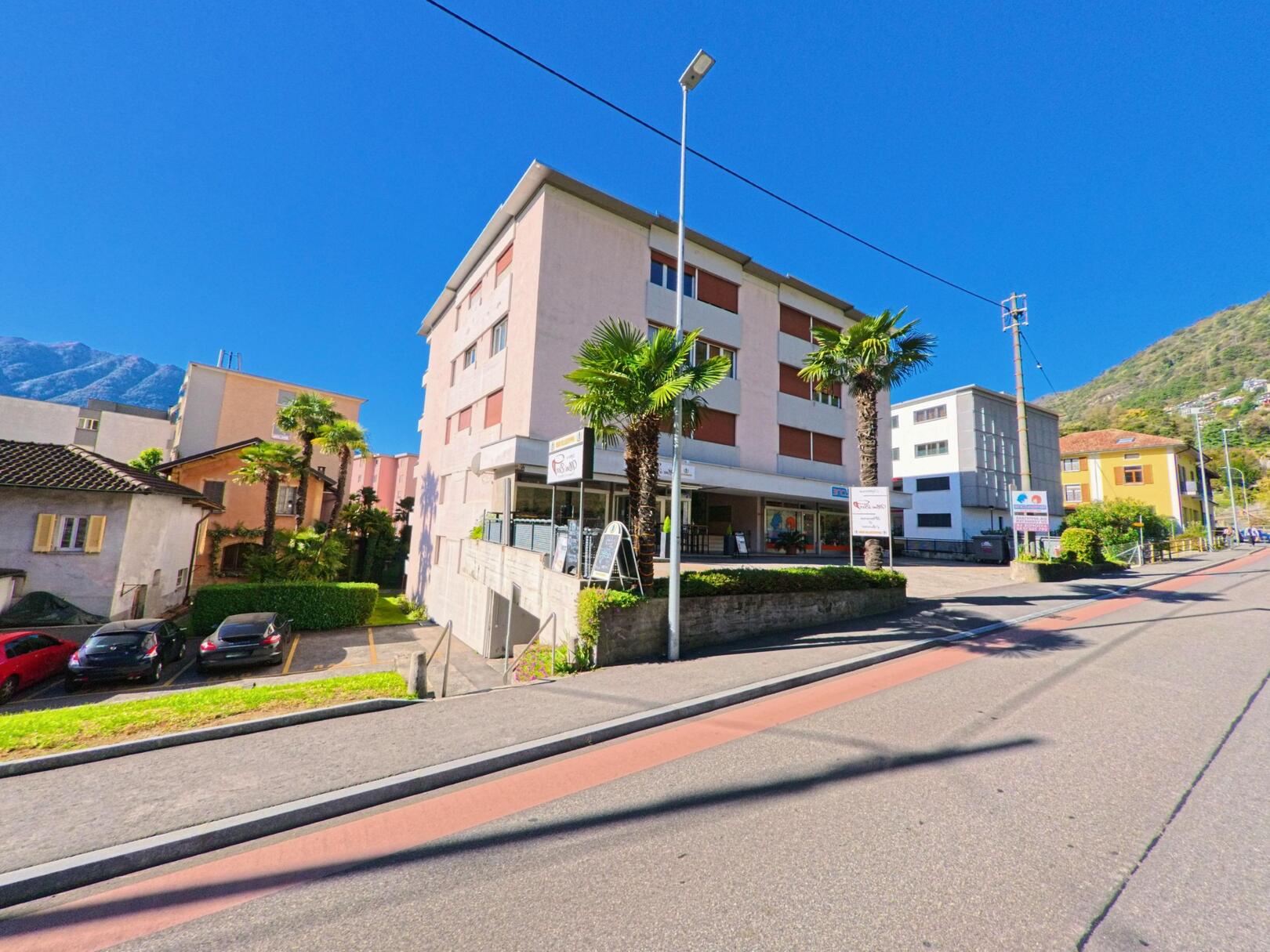 Entlang der Hauptstrasse in Tenero-Contra (6598, Kanton Tessin) stehen moderne Wohn- und Geschäftsgebäude mit Palmen und Blick auf die umliegenden Berge.