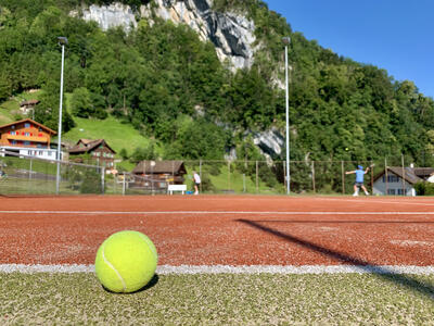 Auf einem Tennisplatz in Gersau (6442, Kanton Schwyz) liegt ein Ball im Vordergrund, waehrend im Hintergrund zwischen Haeusern und Felswand zwei Personen spielen.