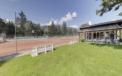 In Grüsch (7214, Kanton Graubünden) liegen mehrere Sandtennisplätze neben einem Clubhaus, umgeben von Grünflächen und Bergen.