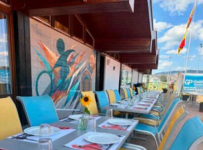 Die Terrasse des Tennisclubs Konolfingen aus Konolfingen (3510, Kanton Bern) ist mit langen gedeckten Tischen, farbigen Stühlen und Sonnenblumen dekoriert, während im Hintergrund die Tennisanlage zu sehen ist.