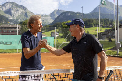 Am Netz eines Sandtennisplatzes gratulieren sich in Unterwasser (9657, Kanton St. Gallen) zwei Tennisspieler nach einem Match vor alpiner Landschaft.