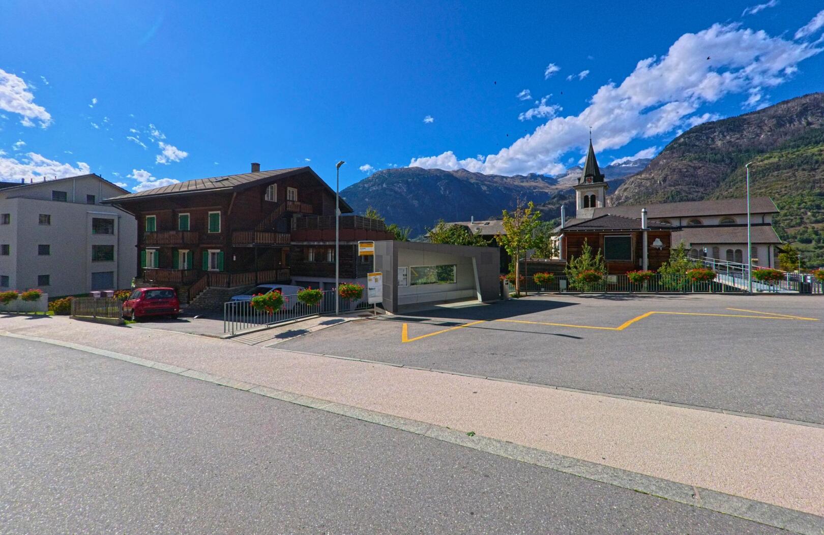 Im Zentrum von Termen (3912, Kanton Wallis) steht eine Bushaltestelle neben traditionellen Holzhäusern und der Kirche vor alpiner Bergkulisse.
