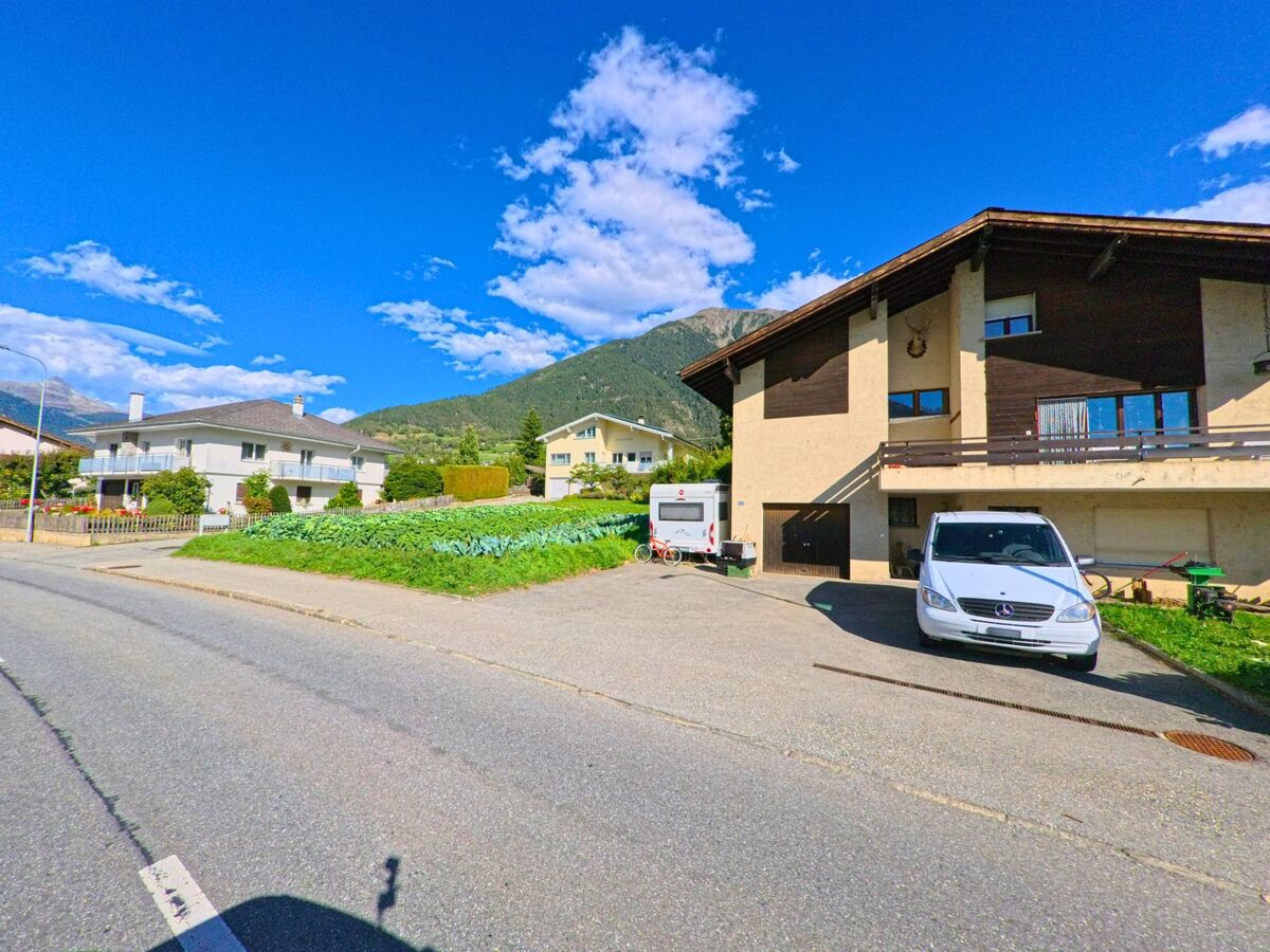 Ein Wohnhaus mit Balkon in Termen (3912, Kanton Wallis) liegt an einer Strasse, im Hintergrund sind grüne Hänge und Berge zu sehen.