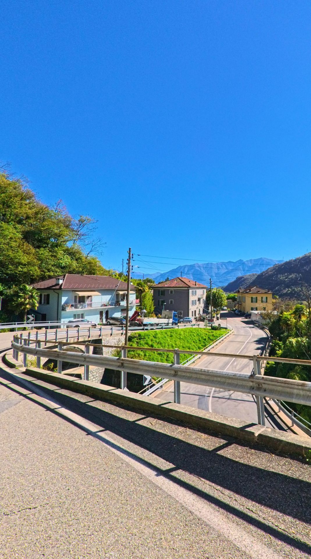 Von einer Brücke in Terre di Pedemonte (6654, Kanton Tessin) bietet sich ein Blick auf mehrere Wohnhäuser und die umliegenden Berge unter wolkenlosem Himmel.