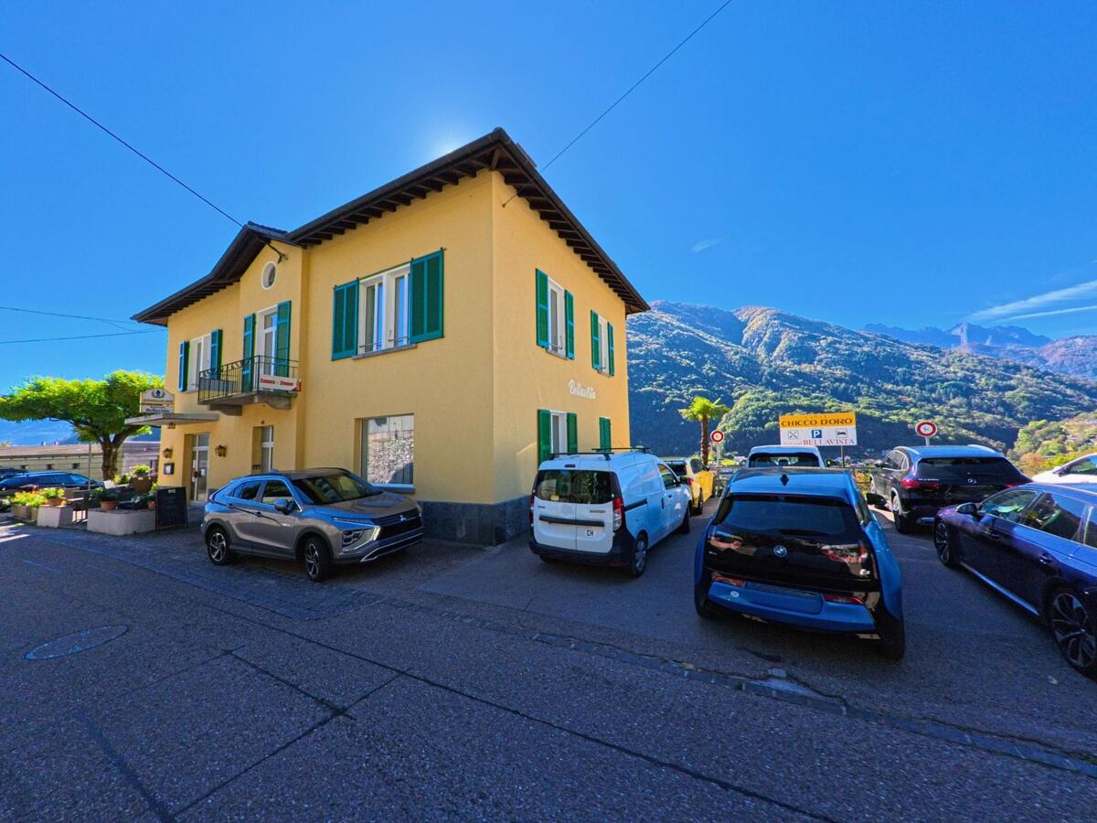 Vor einem gelben Gebäude mit grünen Fensterläden in Terre di Pedemonte (6654, Kanton Tessin) stehen mehrere geparkte Autos, im Hintergrund erheben sich bewaldete Berge.