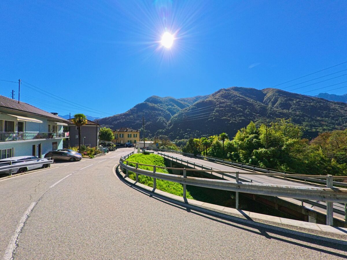 Eine kurvenreiche Strasse in Terre di Pedemonte (6654, Kanton Tessin) führt an Wohnhäusern vorbei, während im Hintergrund bewaldete Hügel unter der hellen Sonne liegen.