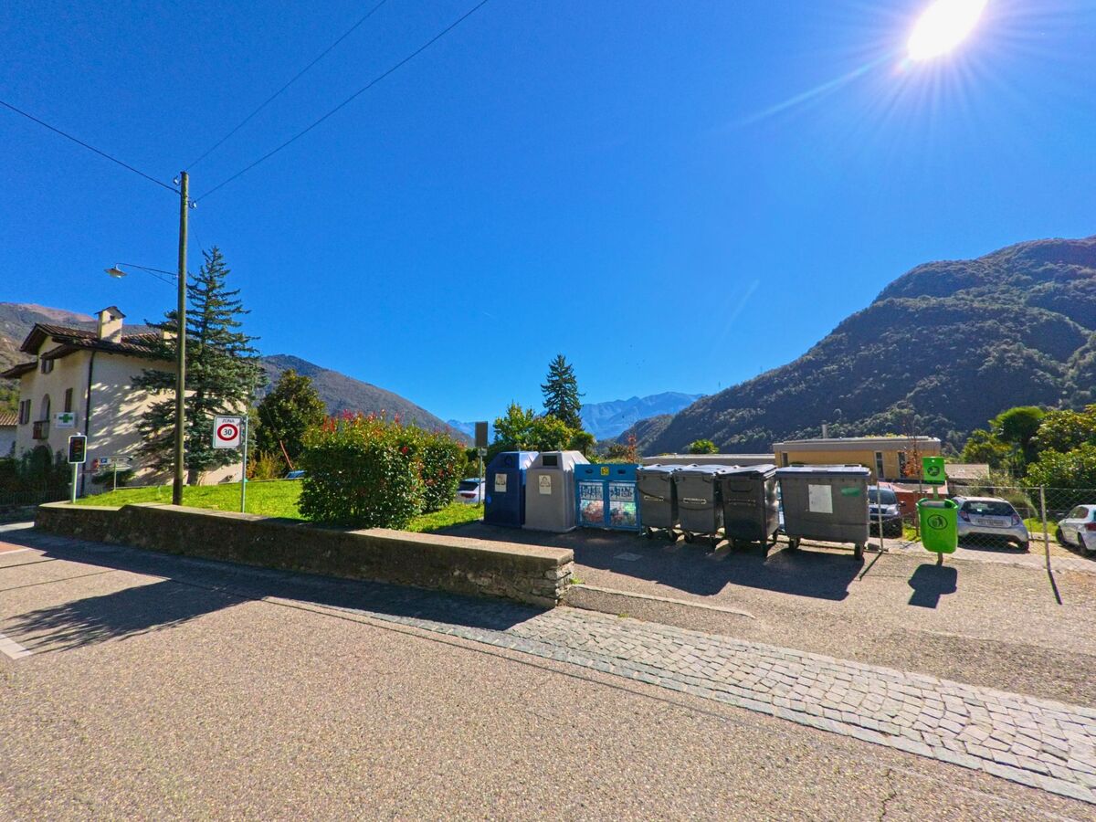 In Terre di Pedemonte (6654, Kanton Tessin) befindet sich eine Recyclingstelle mit Containern und Abfallbehältern vor einer Bergkulisse bei strahlendem Sonnenschein.