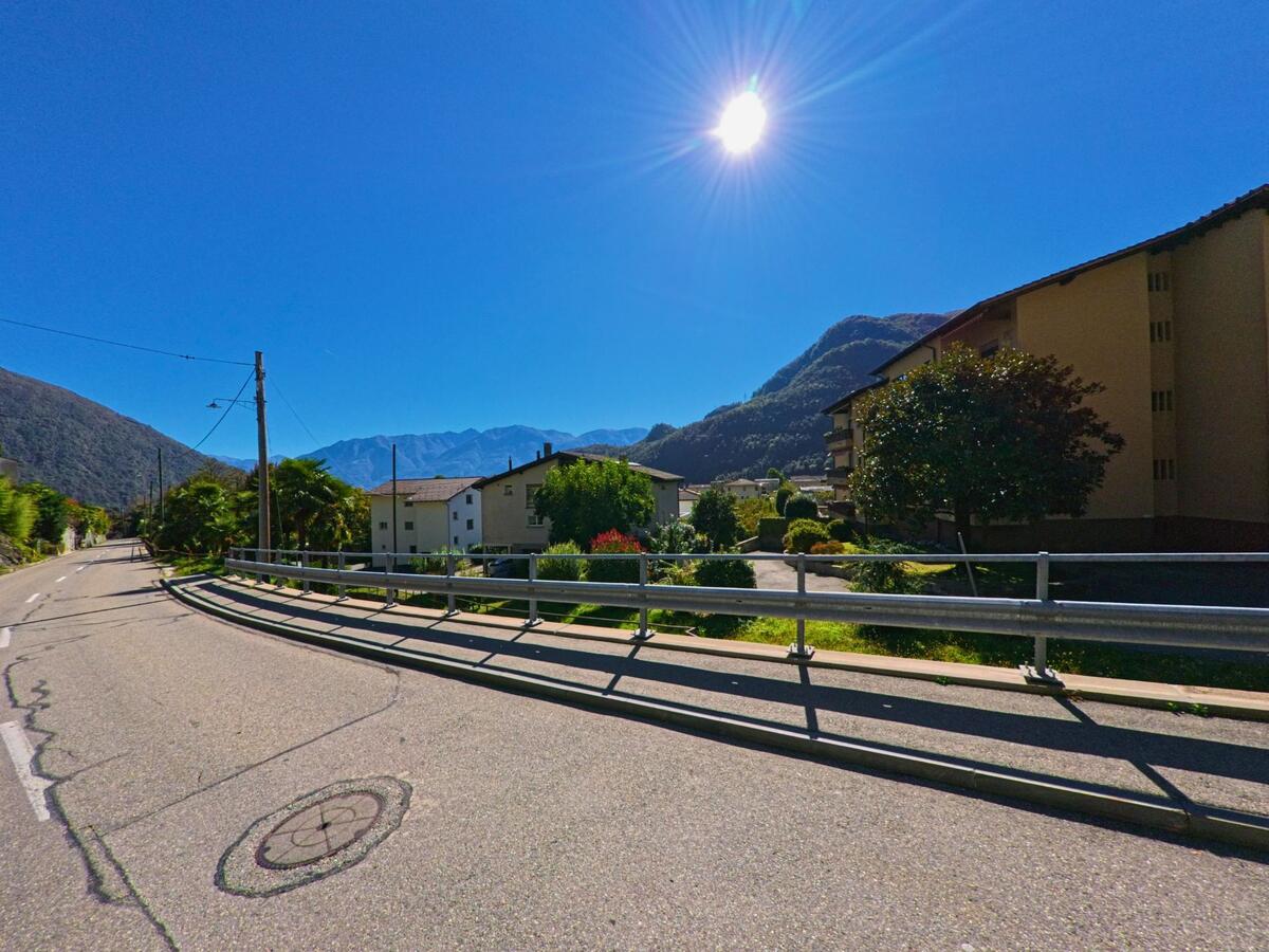 Eine breite Strasse in Terre di Pedemonte (6654, Kanton Tessin) verläuft zwischen Wohnhäusern und Palmen, während die Sonne über den umliegenden Bergen strahlt.