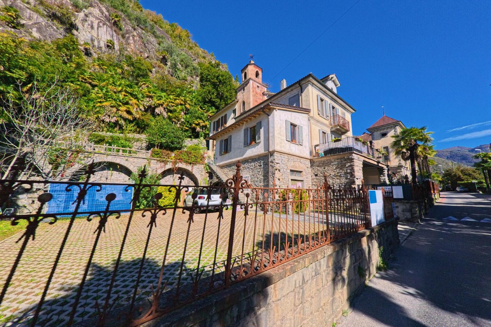Ein historisches Wohnhaus mit Türmchen und steinernen Mauern steht in Terre di Pedemonte (6654, Kanton Tessin) am Fusse eines bewaldeten Hügels unter klarem, blauem Himmel.