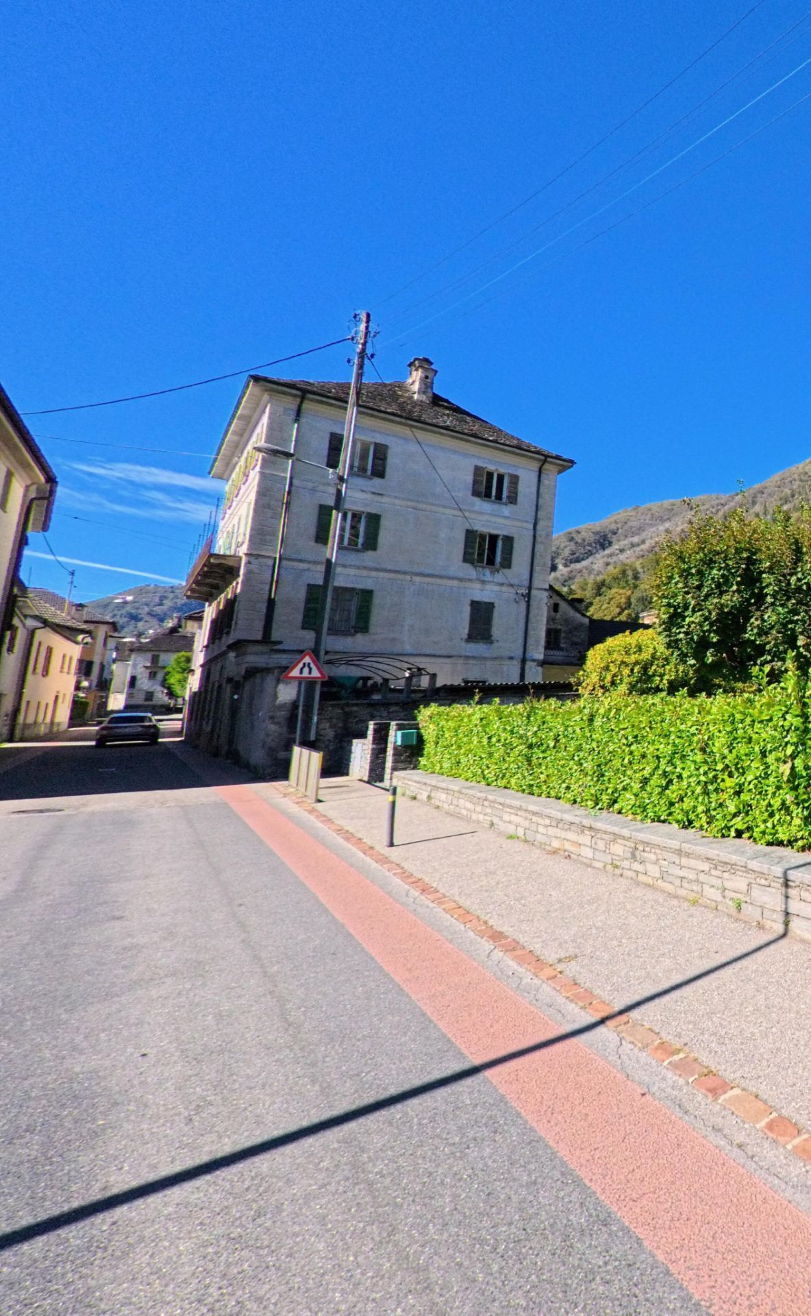 Ein hohes Wohnhaus mit grünen Fensterläden steht an einer schmalen Strasse in Terre di Pedemonte (6654, Kanton Tessin) unter strahlend blauem Himmel.