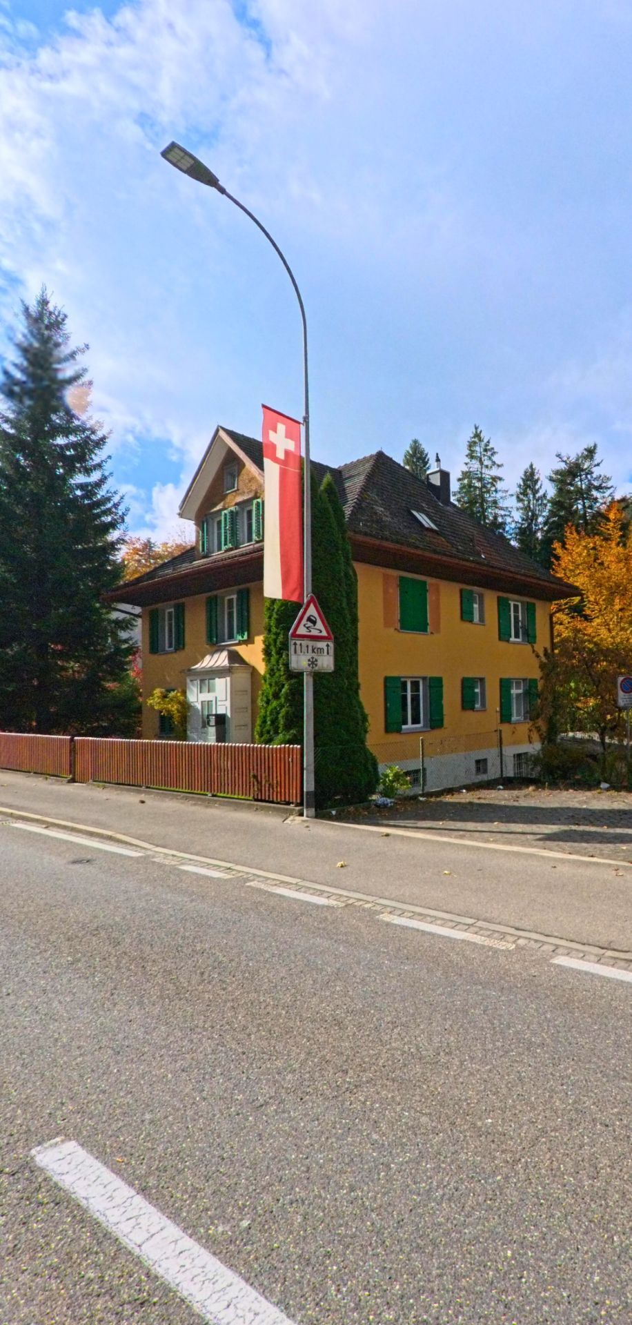 Ein gelbes Haus mit gruenen Fensterlaeden steht in Teufen (9012, Kanton Appenzell Ausserrhoden) an einer Strasse, neben der eine Schweizer Flagge und ein Gefaellewarnschild angebracht sind.