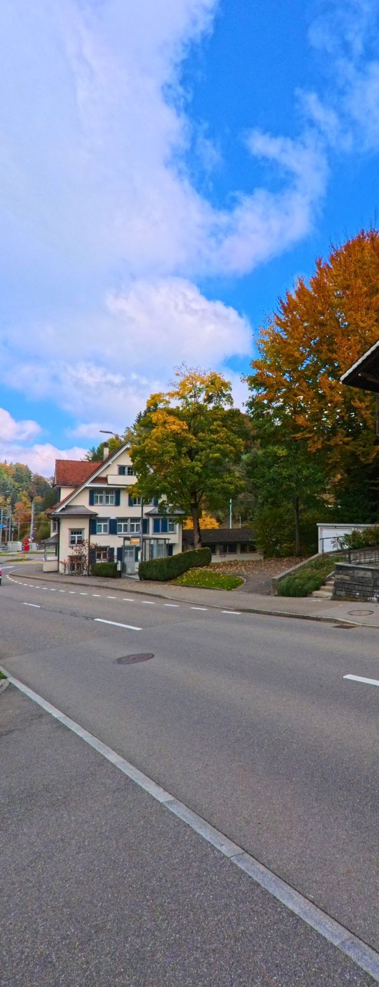 Ein Haus mit blauen Fensterlaeden steht in Teufen (9012, Kanton Appenzell Ausserrhoden) an einer sanft geschwungenen Strasse, flankiert von herbstlichen Baumen und einem kleinen Platz.