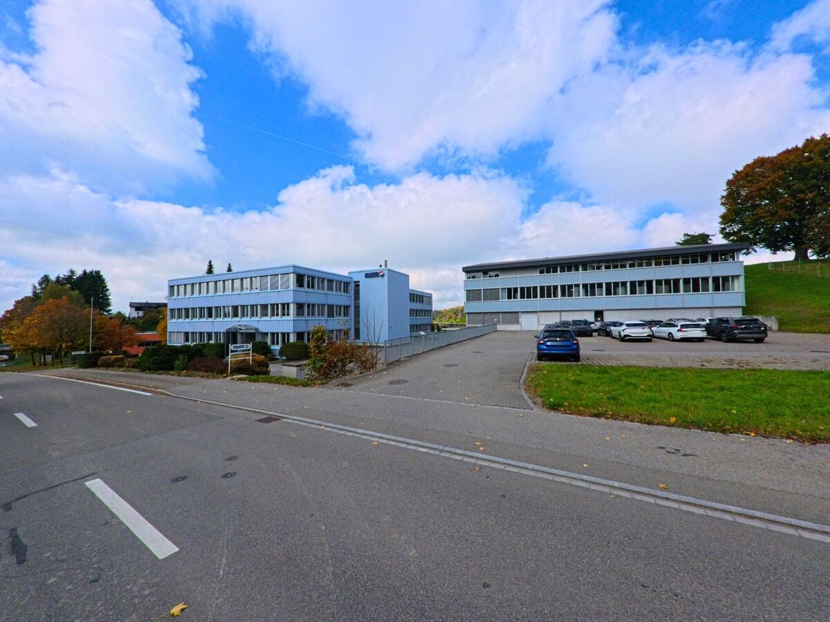 Zwei moderne Buerohaeuser mit grossen Fensterfronten stehen in Teufen (9012, Kanton Appenzell Ausserrhoden) oberhalb einer breiten Strasse, umgeben von Parkplaetzen und herbstlichen Baumpartien.