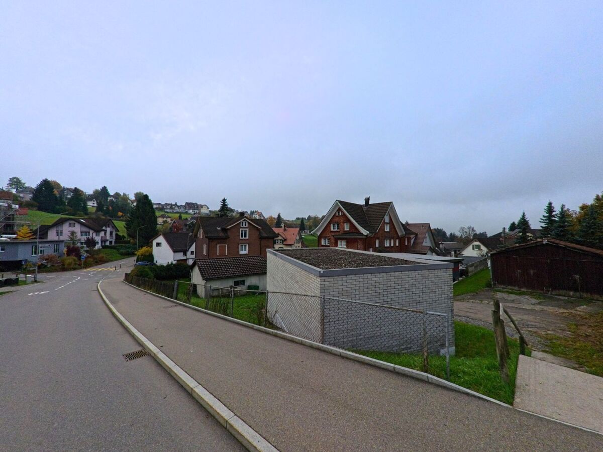 Entlang einer sanft abfallenden Strasse in Teufen (9012, Kanton Appenzell Ausserrhoden) stehen mehrere Wohnhaeuser vor leicht nebliger Huegellandschaft und herbstlichem Baumgruen.