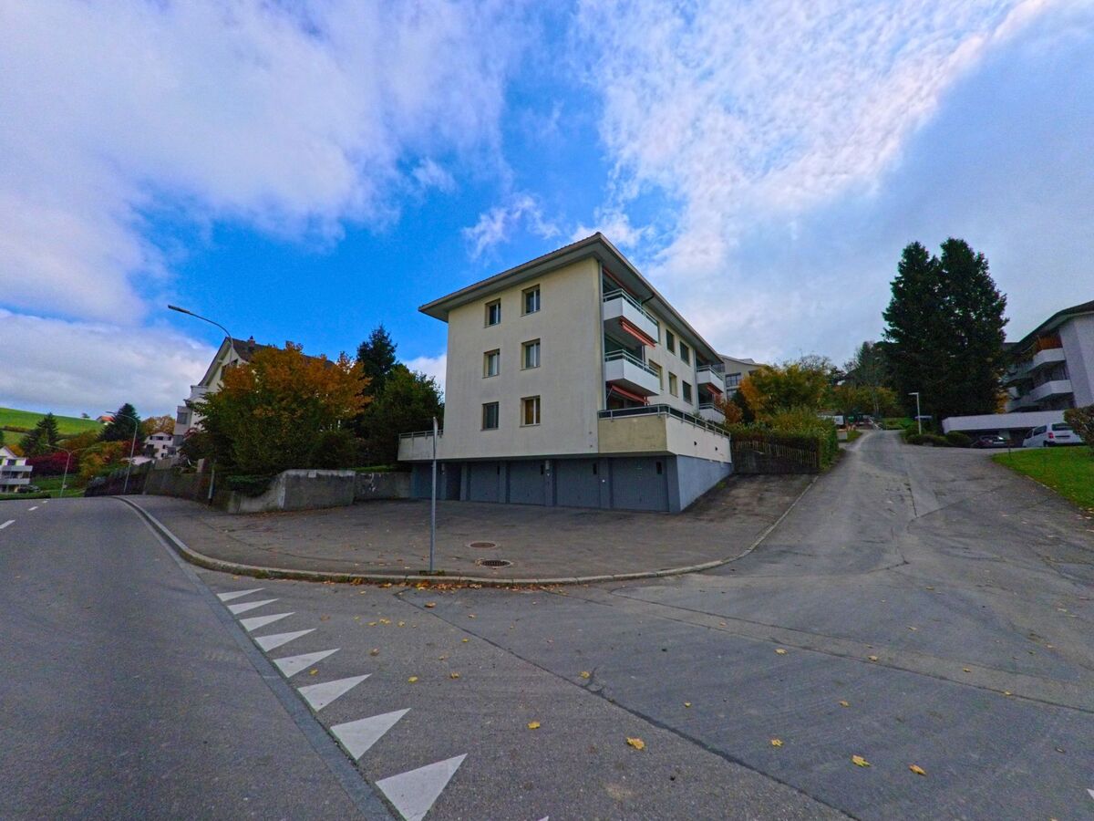 Ein mehrgeschossiges Wohngebaeude mit Balkonen steht in Teufen (9012, Kanton Appenzell Ausserrhoden) an einer abzweigenden Steigung zwischen weiteren Hausern und herbstlichem Gruen.