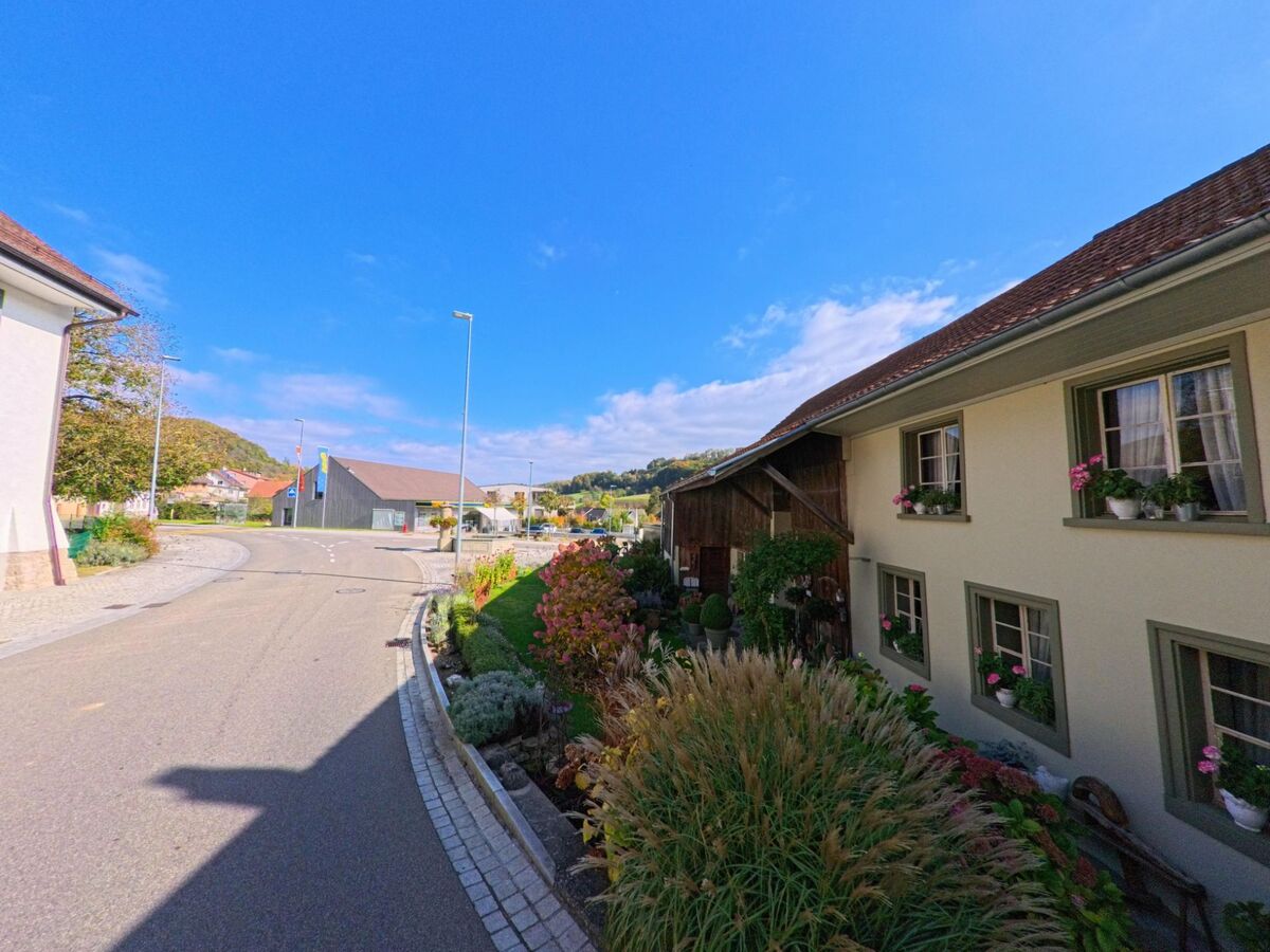 In Thalheim (5112, Kanton Aargau) fuehrt eine ruhige Strasse an einem gepflegten Wohnhaus mit Blumenkisten vorbei, waehrend im Hintergrund eine moderne Holzhalle und der bewaldete Huegel zu sehen sind.