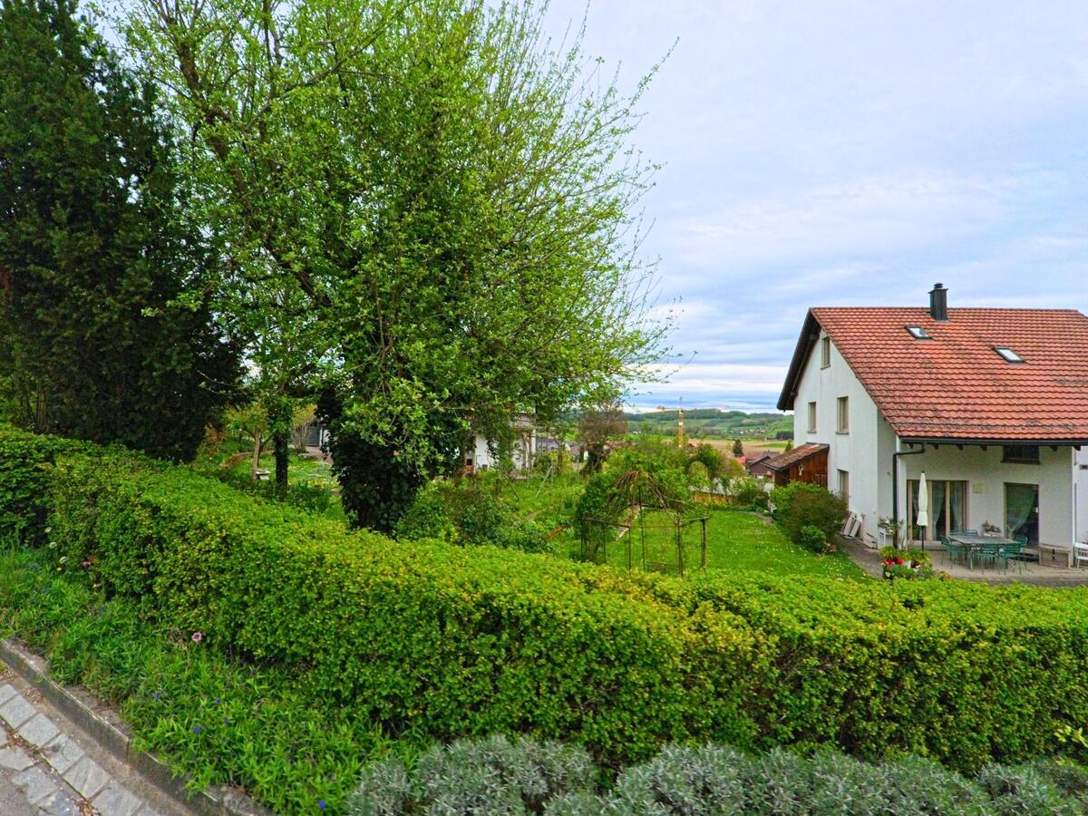 In Thalheim an der Thur (8478, Kanton Zürich) liegt ein Einfamilienhaus mit rotem Ziegeldach hinter gepflegten Hecken und Gärten mit Blick auf die weite Landschaft.