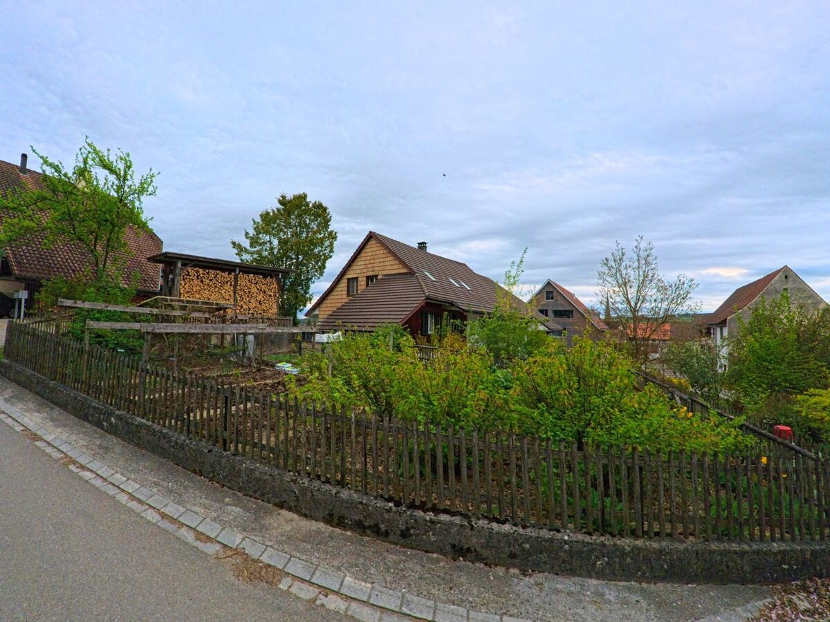  In Thalheim an der Thur (8478, Kanton Zürich) verläuft eine schmale Strasse an einem eingezäunten Gartengrundstück mit Holzlager und mehreren Einfamilienhäusern vorbei.