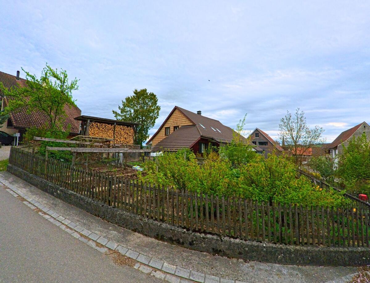 In Thalheim an der Thur (8478, Kanton Zürich) grenzt ein kleiner Hausgarten mit Holzlattenzaun an eine Wohnstrasse mit Einfamilienhäusern und Holzunterständen.