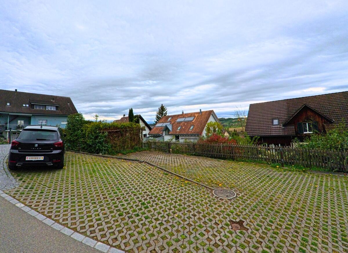 In Thalheim an der Thur (8478, Kanton Zürich) liegt ein Parkplatz mit Rasengittersteinen zwischen Einfamilienhäusern mit Blick auf die leicht hügelige Landschaft.