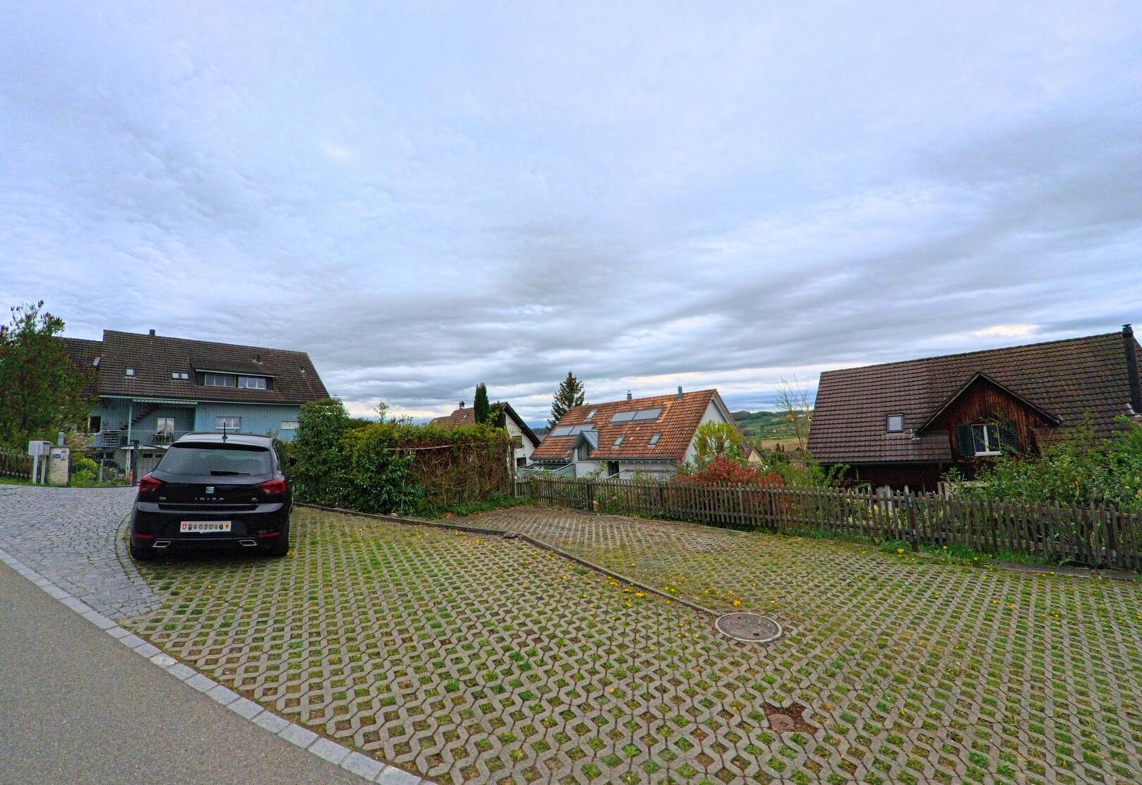 In einem Wohnquartier von Thalheim an der Thur (8478, Kanton Zürich) liegt ein mit Rasengittersteinen gepflasterter Parkplatz vor Einfamilienhäusern mit Blick auf die hügelige Landschaft.