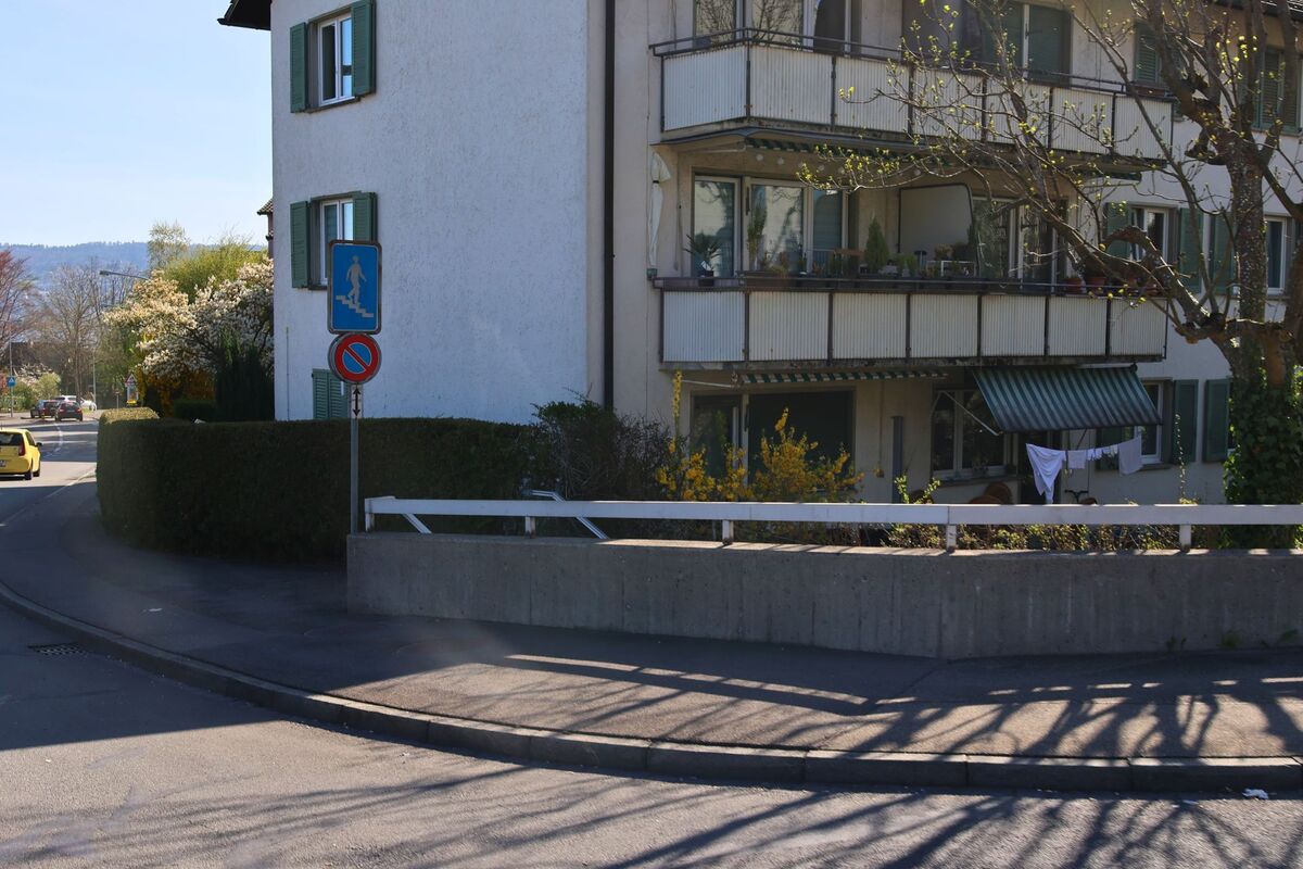 In Thalwil (8800, Kanton Zürich) steht ein Wohnhaus mit grünen Fensterläden und überdachten Balkonen an einer leicht gebogenen Strasse mit Fussgängerschild und Geländer.