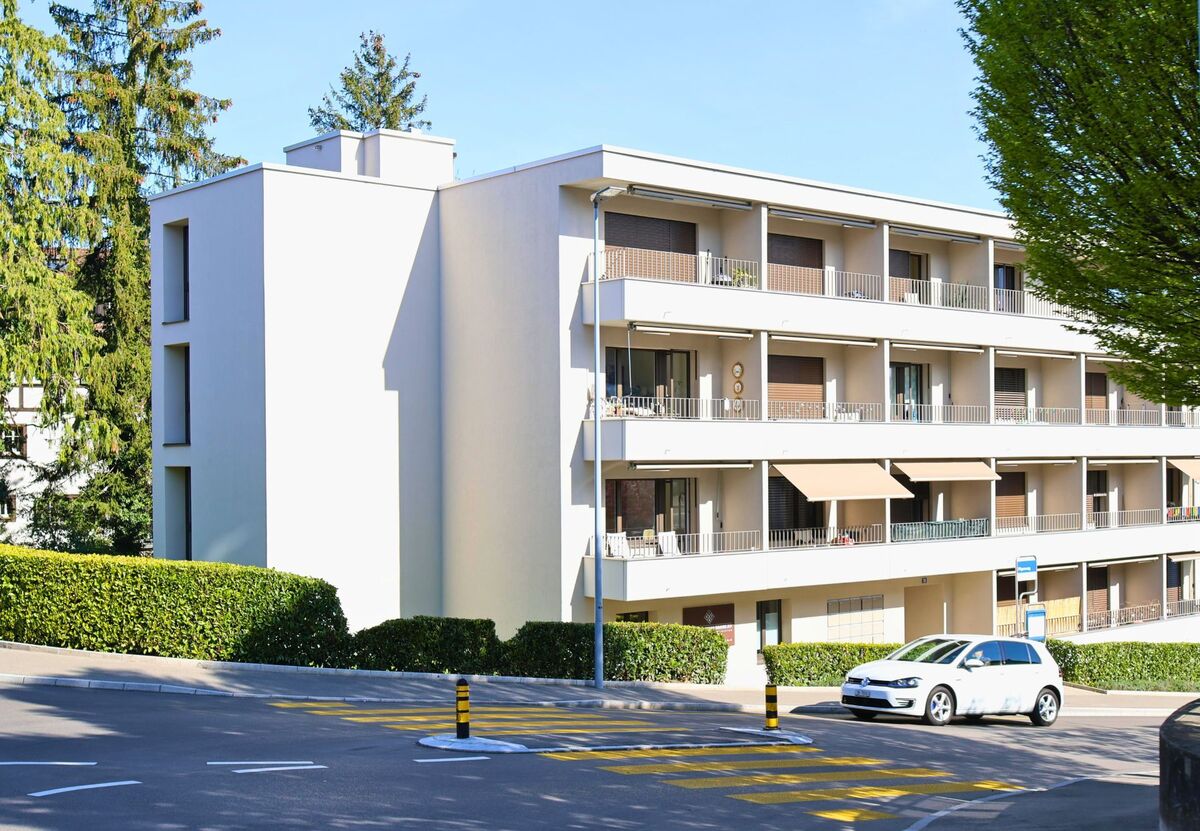 In Thalwil (8800, Kanton Zürich) steht ein modernes Wohnhaus mit grossen Balkonen an einer kurvigen Strasse mit Zebrastreifen und Bushaltestelle.
