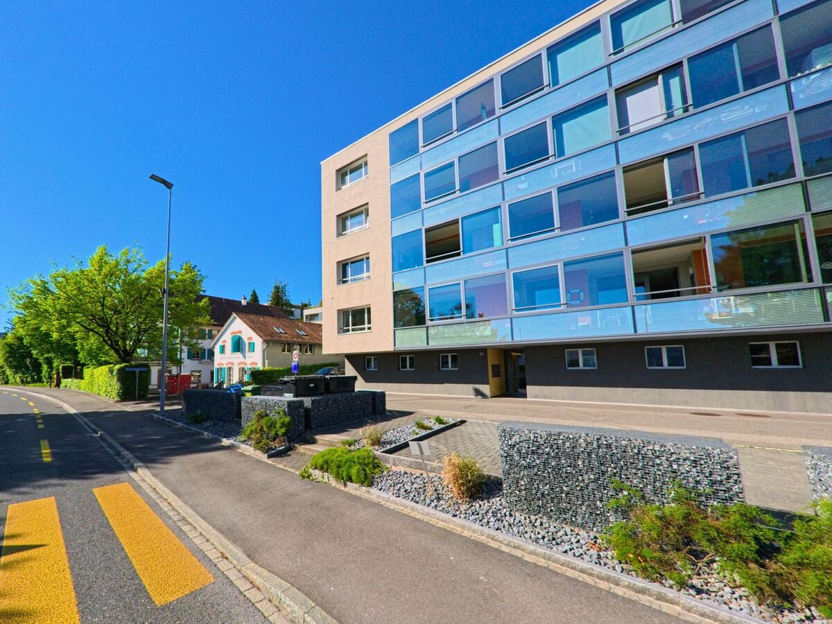 In Thalwil (8803, Kanton Zürich) steht ein modernes Wohngebäude mit grossflächigen Glasfronten direkt an einer Hauptstrasse mit Zebrastreifen und bepflanztem Vorgarten.