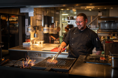 In Lachen (8853, Kanton Schwyz) grillt ein Koch in schwarzer Uniform ein Steak ueber offener Flamme in einer modernen Restaurantkueche.